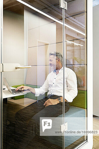 Älterer Geschäftsmann arbeitet am Laptop  während er in einer Kabine im Büro sitzt