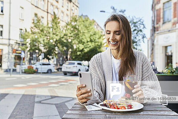 Lächelnde Frau  die ein Mobiltelefon benutzt  während sie in einem Straßencafé frühstückt