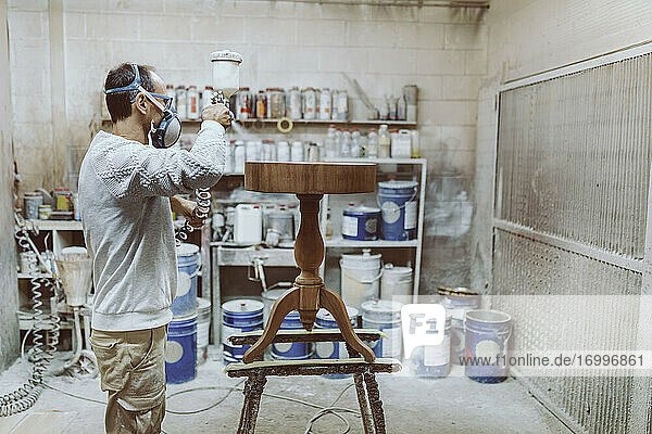 Handwerker mit Gesichtsschutzmaske  der in einer Werkstatt stehend auf einen Tisch sprüht