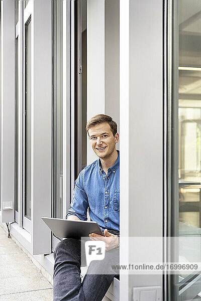 Junger Geschäftsmann  der ein digitales Tablet benutzt  während er an der Bürotür sitzt