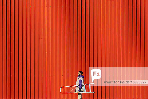 Young man carrying a ladder in front of a red wall