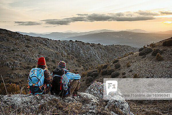 Männliche und weibliche Wanderer genießen den Sonnenuntergang  während sie im Urlaub auf einem Felsenberg sitzen