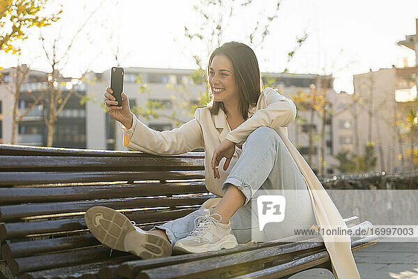 Lächelnde Geschäftsfrau  die ein Selfie mit ihrem Smartphone macht  während sie auf einer Bank sitzt