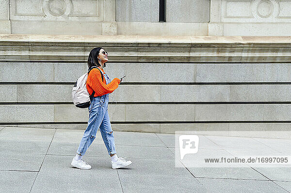Junge Frau mit Rucksack und Mobiltelefon  die beim Gehen auf dem Fußweg wegschaut