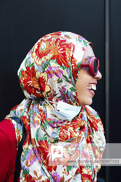 Fröhliche Frau mit geblümtem Hidschab vor schwarzer Wand während der COVID-19