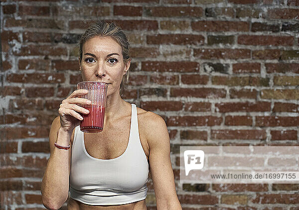 Mature woman drinking fruit smoothie against brick wall in kitchen