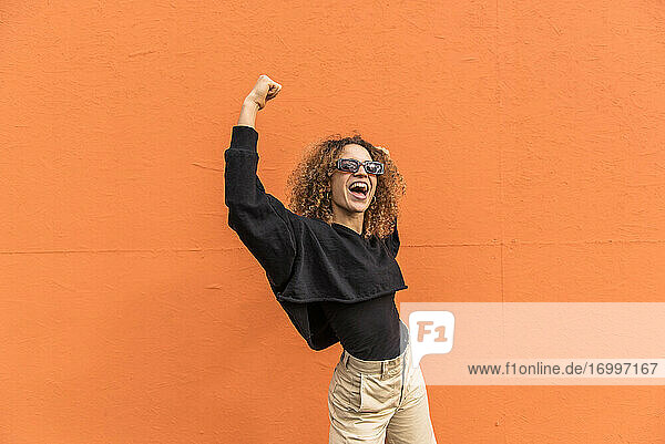 Fröhliche Frau mit erhobenen Armen an einer orangefarbenen Wand stehend
