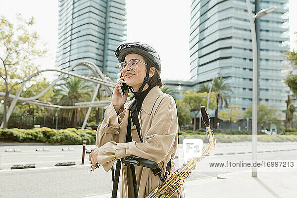 Lächelnde Frau mit Saxophon  die mit ihrem Handy spricht  während sie auf einem Motorroller in der Stadt steht