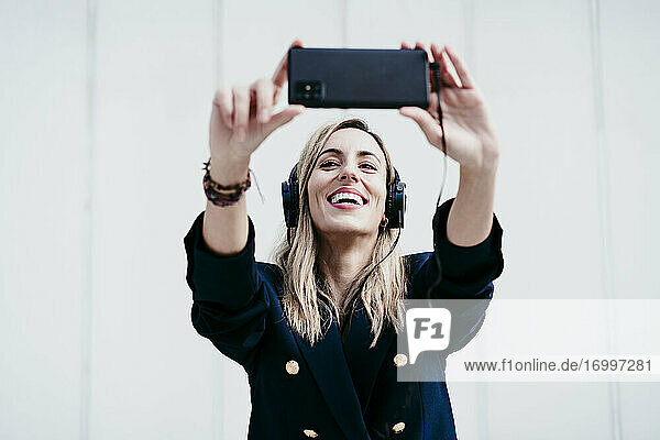 Lächelnde Frau  die ein Selfie mit ihrem Smartphone macht  während sie an der Wand Musik hört