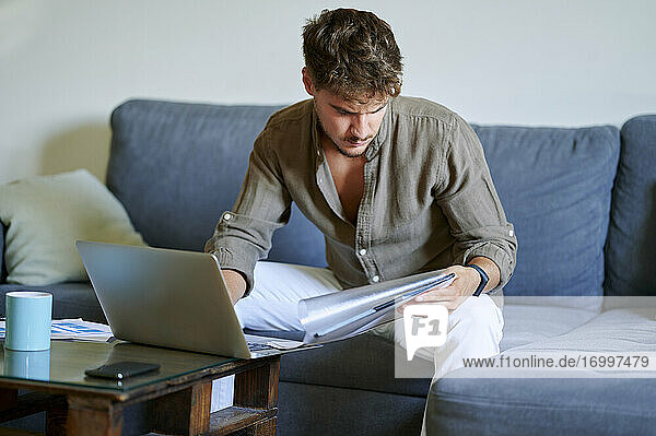 Junger männlicher Freiberufler  der zu Hause auf dem Sofa mit seinem Laptop sitzt und Dokumente liest
