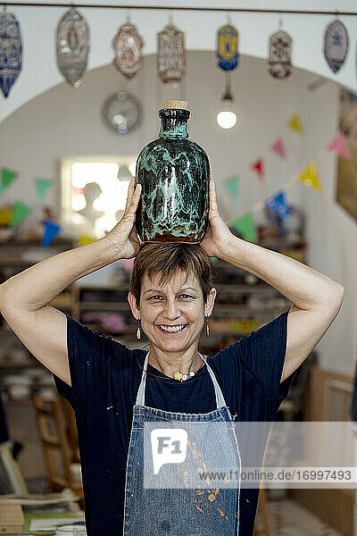 Lächelnde Künstlerin mit türkisfarbener Flasche auf dem Kopf im Keramikladen