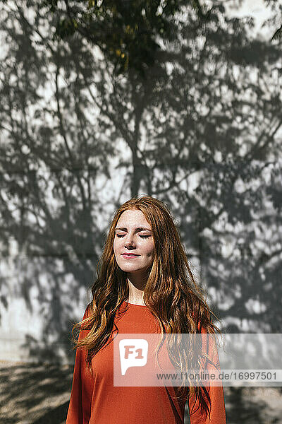 Young redhead woman standing with eyes closed against tree shadow wall