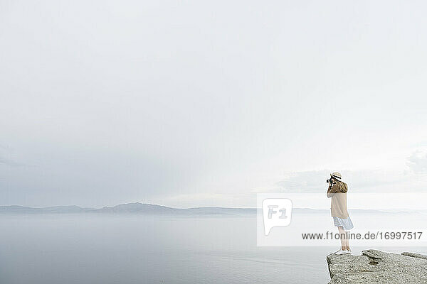 Frau mit Kamera  die auf einem Felsen steht und das Meer fotografiert  Rückansicht