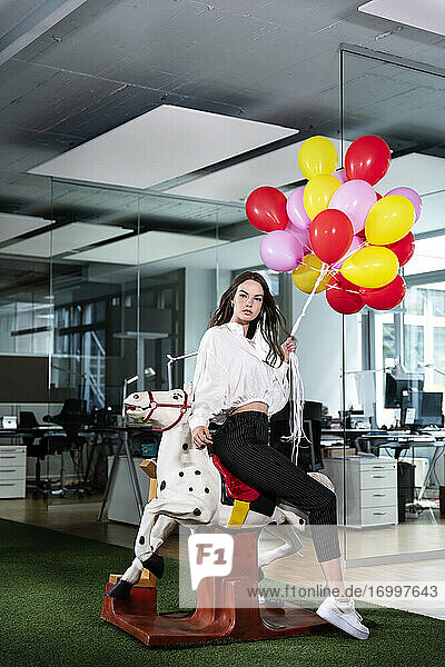 Junge Geschäftsfrau mit buntem Ballon sitzt auf Schaukelpferd im Büro