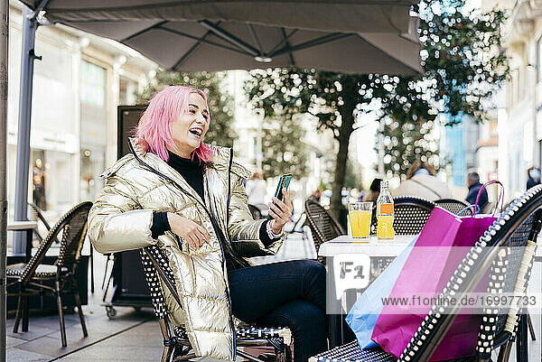 Frau mit rosa Haaren  die ein Mobiltelefon benutzt  während sie mit Einkaufstaschen in einem Straßencafé sitzt