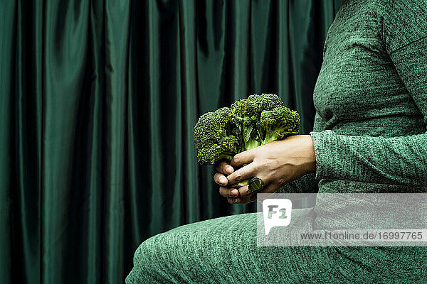 Mittelteil einer Frau  die Brokkoli hält und vor einem grünen Vorhang sitzt