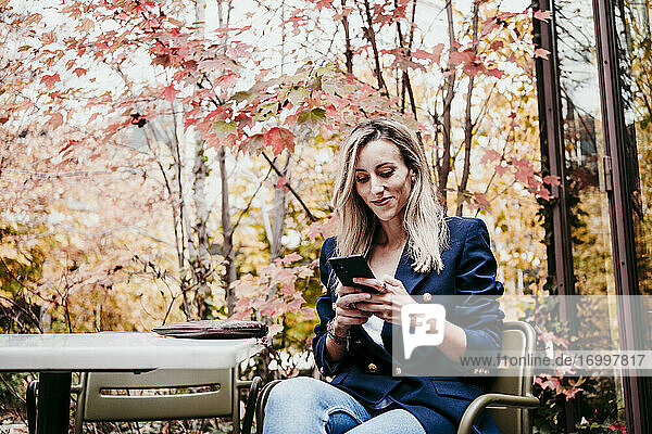Blonde Frau im mittleren Erwachsenenalter  die ein Smartphone benutzt  während sie auf der Terrasse vor den Herbstbäumen sitzt