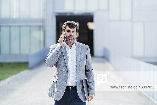 Männlicher Berufstätiger beim Gehen und Telefonieren vor einem Bürogebäude
