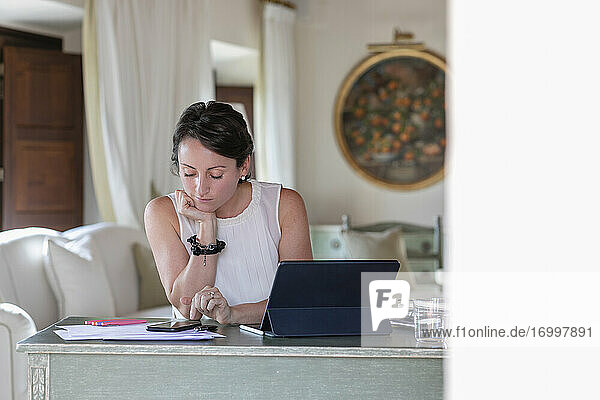 Geschäftsfrau mit Hand am Kinn  die bei der Arbeit zu Hause ein Smartphone benutzt