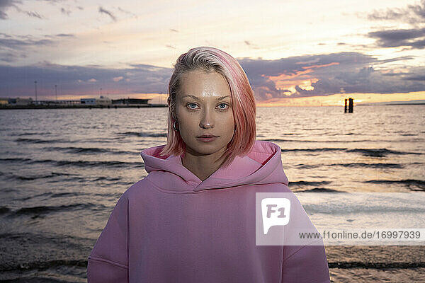 Porträt einer jungen Frau mit rosa Haaren und rosa Kapuzenshirt mit Meer bei Sonnenuntergang im Hintergrund