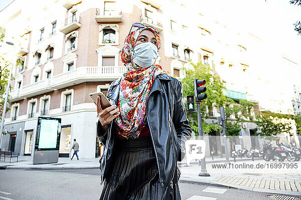 Muslimische Frau im Hidschab mit Handy in der Hand  die während des COVID-19 auf einer Straße in der Stadt geht