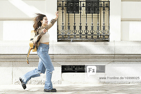 Fröhliche Musikerin mit ausgestreckten Armen  die auf dem Fußweg neben einem Gebäude in der Stadt läuft
