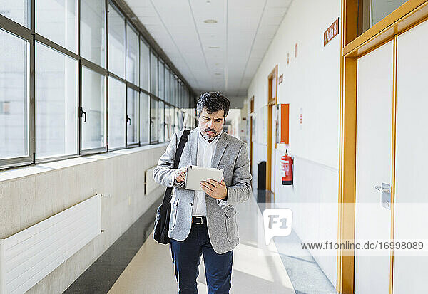 Professor benutzt digitales Tablet  während er durch den Korridor einer Universität geht