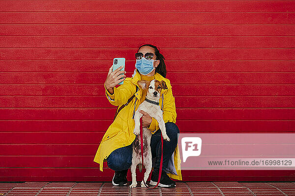 Frau mit Gesichtsmaske und Hund  die Smartphone-Selfies vor einer roten Wand macht