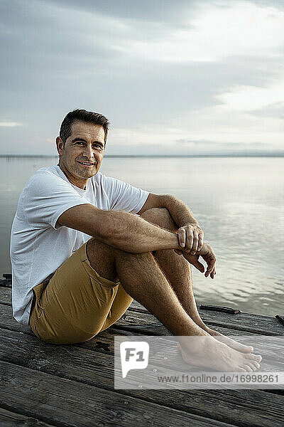 Lächelnder reifer Mann sitzt auf einem Steg am See