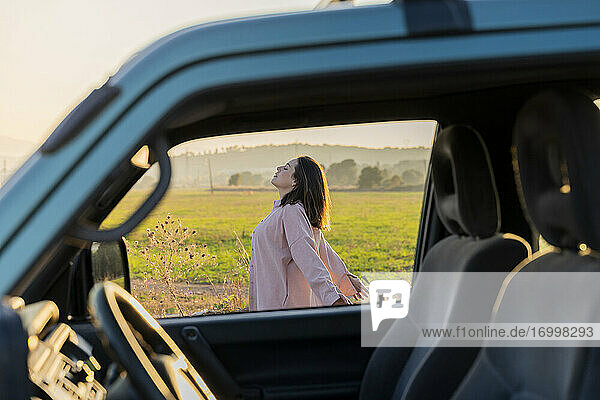Junge Frau  die vom Autofenster aus die Landschaft bei Sonnenuntergang genießt