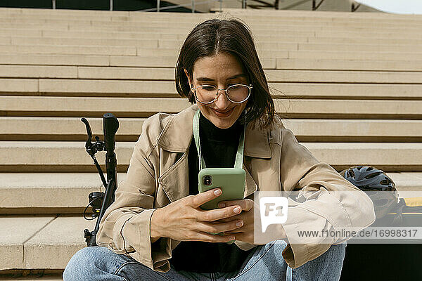 Frau lächelt  während sie ein Mobiltelefon benutzt  während sie mit einem elektrischen Tretroller auf einer Treppe sitzt