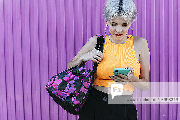 Blonde Frau mit Tasche  die ihr Smartphone vor einer lila Wand benutzt