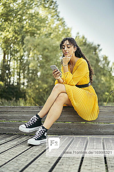 Junge Frau in gelbem Kleid und schwarzen Turnschuhen  die auf einer Holzpromenade ihr Smartphone benutzt
