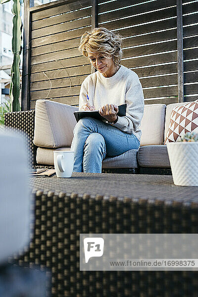 Reife Frau schreibt in Tagebuch auf dem Sofa im Dachgarten sitzend