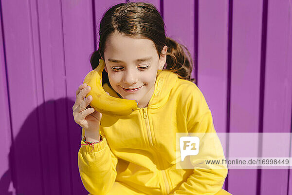 Portrait of smiling girl dressed in yellow holding banana in front of purple background