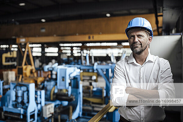 Lächelnder männlicher Unternehmer mit verschränkten Armen in einer Fabrik stehend