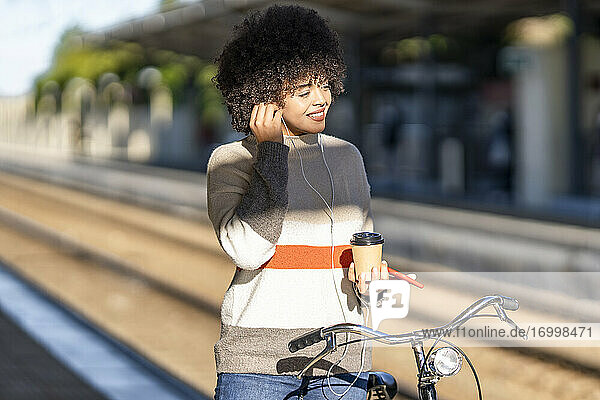 Lächelnde junge Frau mit Fahrrad  die einen wiederverwendbaren Becher in der Hand hält  während sie am Bahnhof Musik genießt
