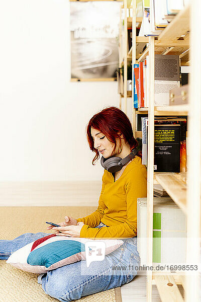 Junge Frau mit Kopfhörern  die ein Mobiltelefon benutzt  während sie zu Hause am Bücherregal sitzt