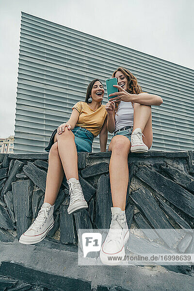 Lächelnde Freundinnen  die ein Mobiltelefon benutzen  während sie an der Wand sitzen
