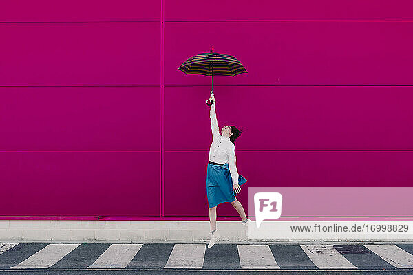 Junge Frau springt mit Regenschirm vor einer rosa Wand