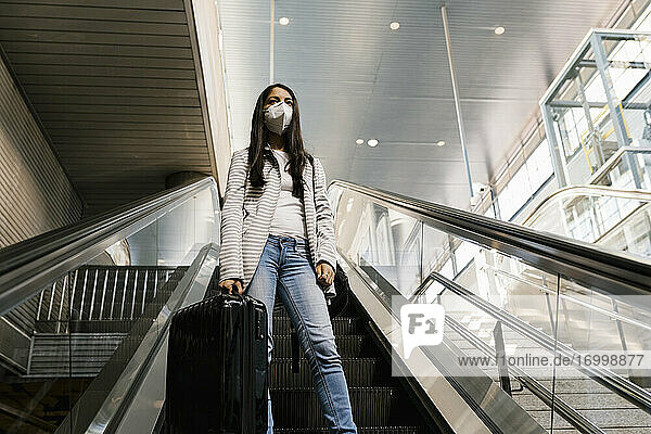 Frau mit Gesichtsmaske  während sie mit Gepäck auf einer Rolltreppe in einer U-Bahn-Station steht