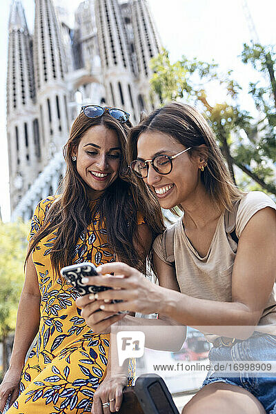 Lächelnde Frauen  die ein Mobiltelefon benutzen  während sie vor der Sagrada Familia in Barcelona  Katalonien  Spanien sitzen