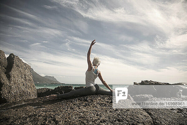 Frau mit gespreizten Beinen und erhobener Hand übt Yoga auf einer Felsformation