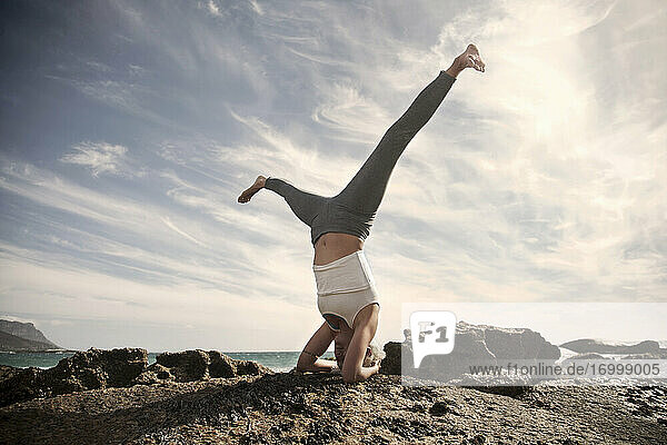 Sorglose Frau übt Sirsasana mit gespreizten Beinen auf einer Felsformation am Strand gegen den Himmel