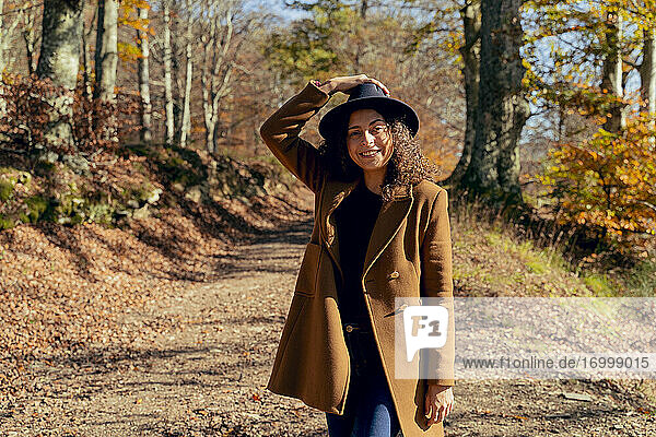 Lächelnde Frau mit Hut und Jacke auf einem Waldweg stehend