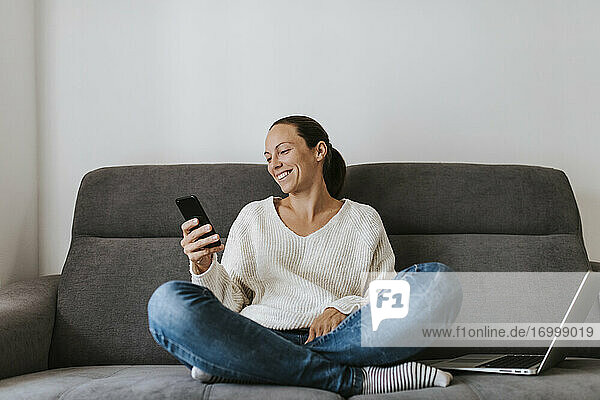 Lächelnde Frau  die ein Smartphone benutzt  während sie mit einem Laptop auf dem Sofa sitzt