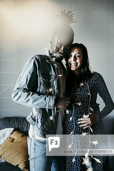 Happy multi-ethnic couple with star shaped lighting equipment standing against wall at home