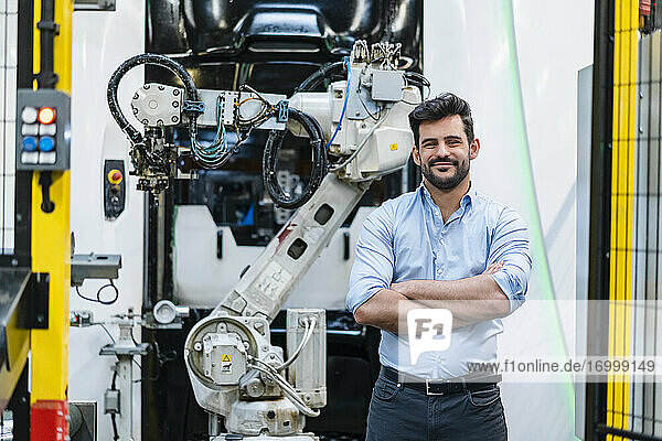 Lächelnder männlicher Unternehmer mit verschränkten Armen  der vor einem Roboterarm in der Industrie steht