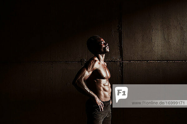 Nachdenklicher Sportler mit nackter Brust und Hand auf der Hüfte  der bei Sonnenuntergang an einer grauen Wand steht