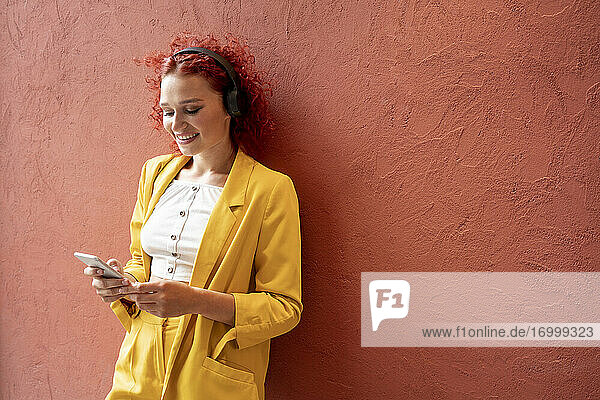Junge Frau in gelbem Anzug lehnt an einer roten Wand  trägt Kopfhörer und benutzt ein Smartphone